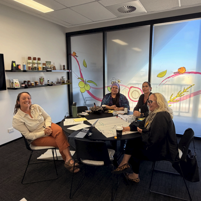 Four women seated around a round table in a meeting room decorated with native plant artwork on the frosted windows.
